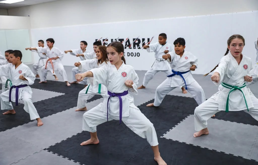 Group of children in white karate uniforms practicing martial arts in a dojo studio with black mat flooring.
